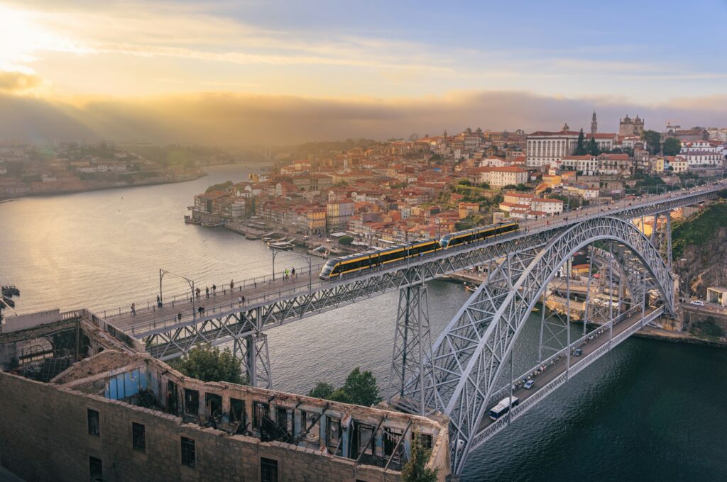 Drone shot of the train bridge in Porto crossing the river. Pedestrains walk along the train line. A modern train crossing the bridge as the sun sets. Showcasing how to travel portugal by train.