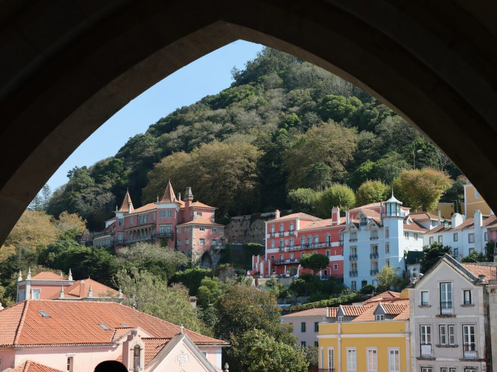 looking through some arches at the beautiful small towns in portugal called sintra. a lush hill with trees and pink buildings.