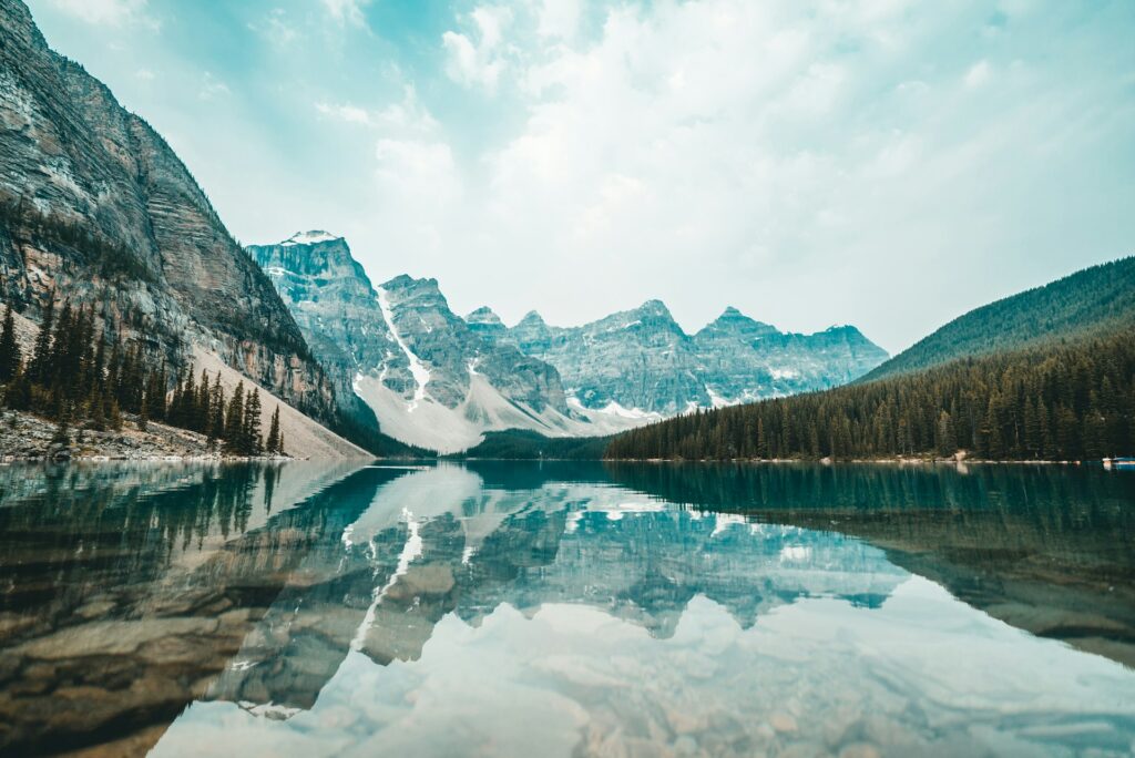 shoreside view of lake luise in western canada. tall mopuntains reflecting off the water and a hill on the right covered in pine trees. a must see on your canada bucket list.