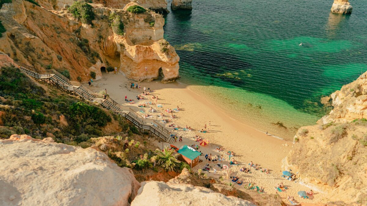 Overview of a beautiful beach in Lagos, Portugal, with stairs leading down a cliff. The sandy shore is filled with people in colorful swimwear, lounging under umbrellas, and enjoying the crystal-clear blue waters. Perfect spot to explore when considering best places to stay in Lagos Portugal.